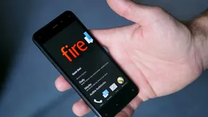 Hand holding Amazon Fire Phone displaying Fire OS home screen with Firefly, Mayday, and Amazon apps interface